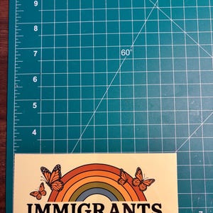 May include: A rectangular sticker with a cream background features a rainbow design with orange, yellow, and green arches, and orange butterflies. The text "IMMIGRANTS MAKE AMERICA GREAT" is printed in bold, black letters.