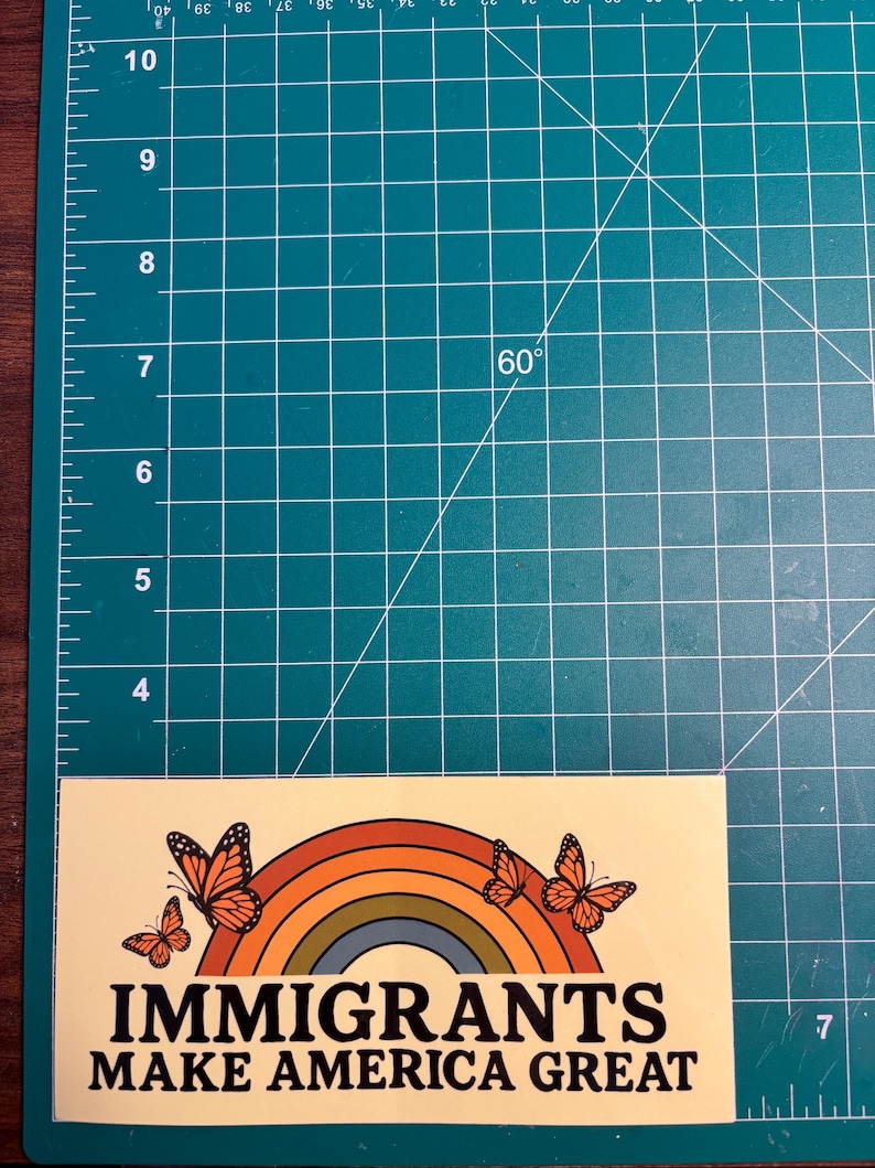 May include: A rectangular sticker with a cream background features a rainbow design with orange, yellow, and green arches, and orange butterflies. The text "IMMIGRANTS MAKE AMERICA GREAT" is printed in bold, black letters.
