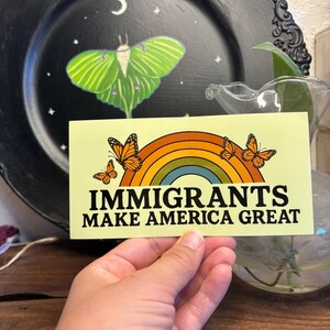 May include: A rectangular sticker with the text "IMMIGRANTS MAKE AMERICA GREAT" above a rainbow and three orange butterflies. The sticker is held in a hand, with a black plate featuring a green moth design in the background.