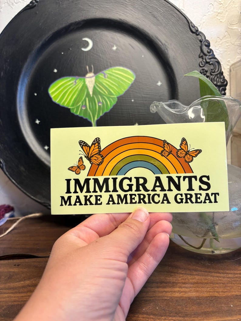 May include: A rectangular sticker with the text "IMMIGRANTS MAKE AMERICA GREAT" above a rainbow and three orange butterflies. The sticker is held in a hand, with a black plate featuring a green moth design in the background.