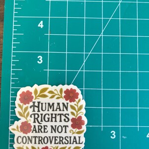 May include: A sticker with the text "HUMAN RIGHTS ARE NOT CONTROVERSIAL" in black, surrounded by a floral design with red flowers and gold leaves. The sticker is on a green cutting mat with inch measurements.