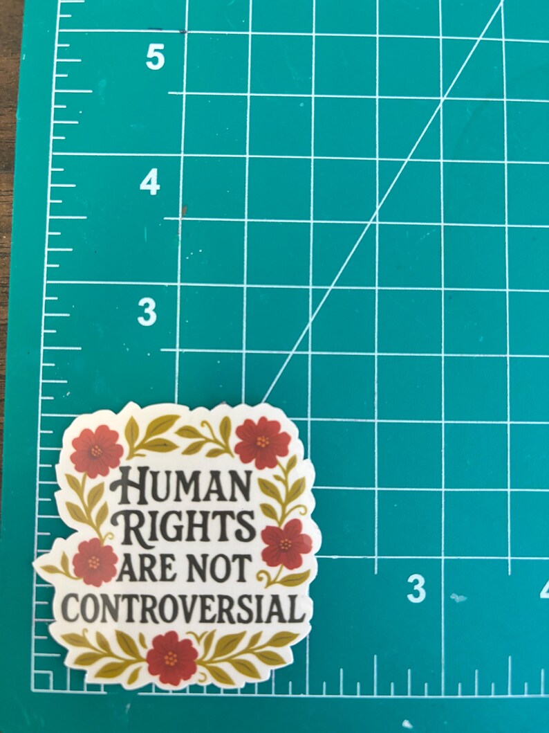 May include: A sticker with the text "HUMAN RIGHTS ARE NOT CONTROVERSIAL" in black, surrounded by a floral design with red flowers and gold leaves. The sticker is on a green cutting mat with inch measurements.