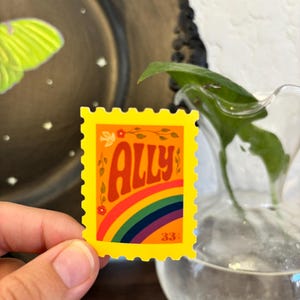 May include: A yellow sticker with a rainbow and the word "ALLY" in orange script. The sticker has a postage stamp design with a floral accent and the number "33".