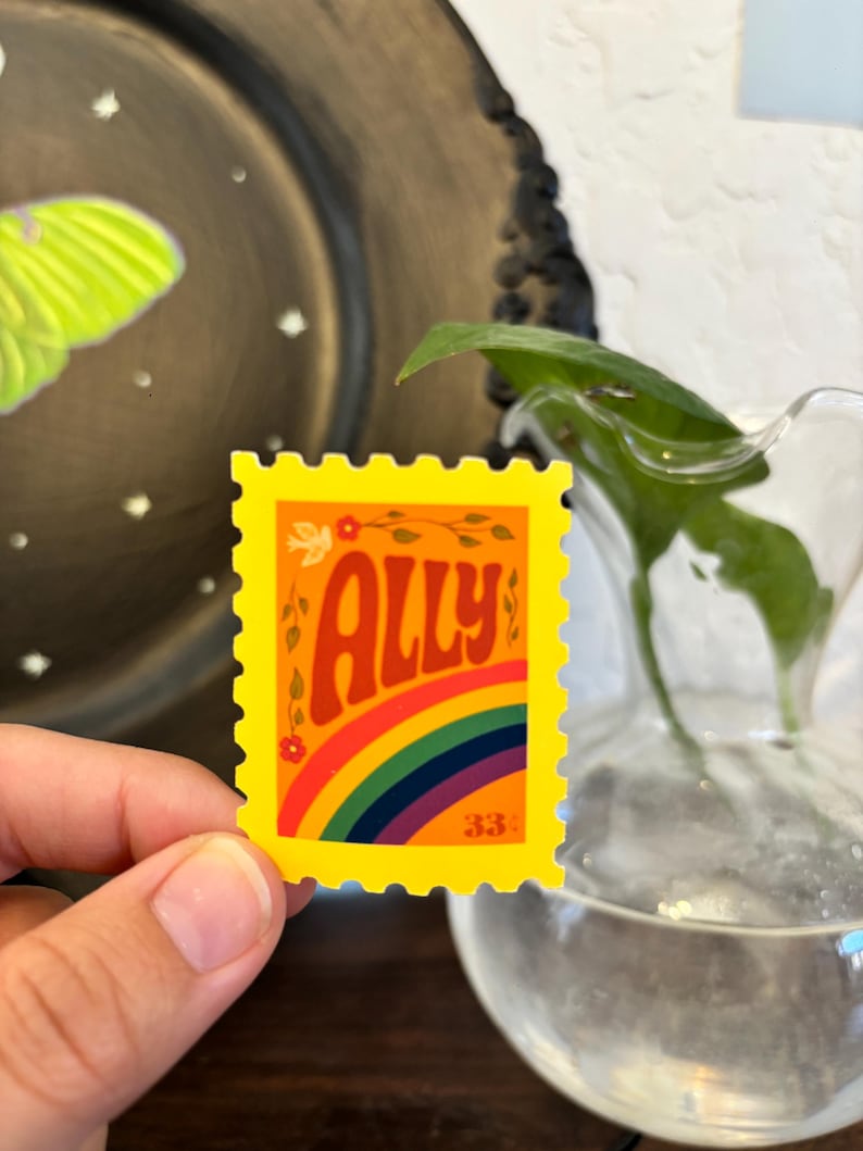 May include: A yellow sticker with a rainbow and the word "ALLY" in orange script. The sticker has a postage stamp design with a floral accent and the number "33".
