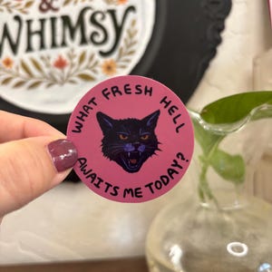 May include: A round sticker with the text "WHAT FRESH HELL AWAITS ME TODAY?" encircling a black cat face on a pink background. The sticker is held in a hand, with a blurred background including a decorative frame and a glass vase.