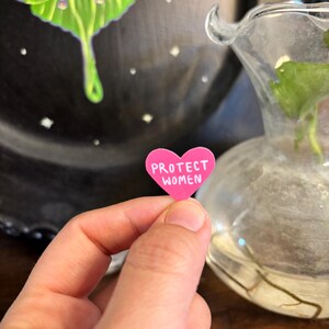 May include: A small, pink heart-shaped sticker with the words "PROTECT WOMEN" in white lettering. The sticker is held in a person's hand, with a blurred background featuring a decorative plate and a glass vase.