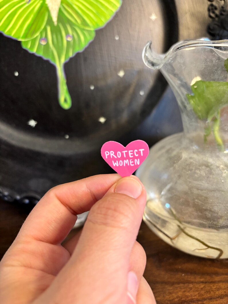 May include: A small, pink heart-shaped sticker with the words "PROTECT WOMEN" in white lettering. The sticker is held in a person's hand, with a blurred background featuring a decorative plate and a glass vase.