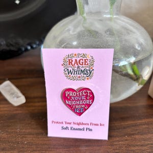 May include: A pink soft enamel pin in the shape of a heart with the text "PROTECT YOUR NEIGHBORS FROM ICE". The pin is on a pink card with the "RAGE & WHIMSY" logo and the text "Protect Your Neighbors From Ice Soft Enamel Pin".