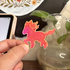 May include: A sticker of a stylized horse with a fiery mane and tail, in shades of pink and orange. The horse is in a running pose, with its legs extended. The sticker is held by a hand.