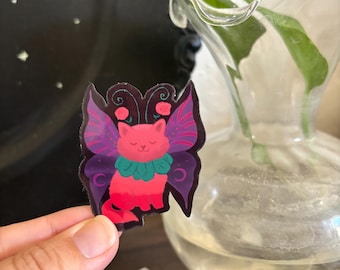May include: A colorful sticker featuring a cat with butterfly wings. The cat is pink and red with a green collar. The wings are purple with pink accents. The sticker is held in a hand.