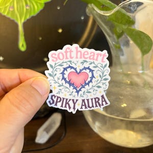 May include: A sticker with the words "soft heart" and "spiky aura". The sticker features a pink heart with blue thorns, surrounded by green leaves. The sticker is held in a hand.