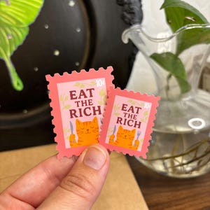 a stamp shaped sticker featuring an orange tabby cat holding up a fork and knife. the text reads “eat the rich.”