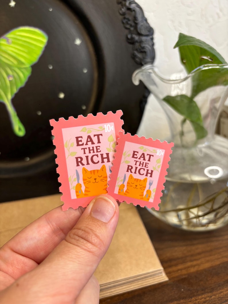 a stamp shaped sticker featuring an orange tabby cat holding up a fork and knife. the text reads “eat the rich.”