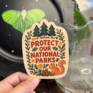 May include: A sticker with the text "PROTECT OUR NATIONAL PARKS" in brown letters. The sticker features illustrations of pine trees, a squirrel, flowers, and acorns, surrounded by a decorative border. The sticker is held in front of a black plate with a moth design.