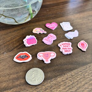 May include: A collection of colorful feminist-themed stickers with various designs and text, including lips with "FEMINIST" and a uterus with "fuck off". A US quarter is included for size reference.