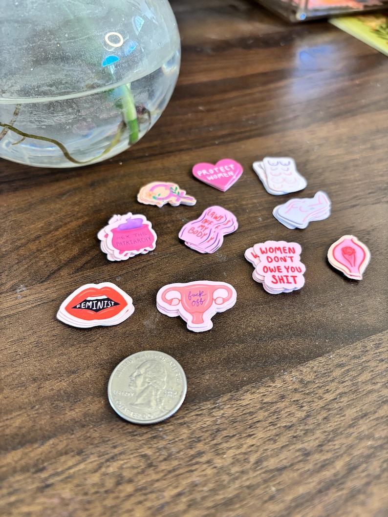 May include: A collection of colorful feminist-themed stickers with various designs and text, including lips with "FEMINIST" and a uterus with "fuck off". A US quarter is included for size reference.