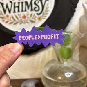 May include: A purple sticker with a jagged edge, displaying the text "PEOPLE>PROFIT" in pink. The sticker is held in front of a blurred background featuring a decorative plate with the words "RAGE & WHIMSY".