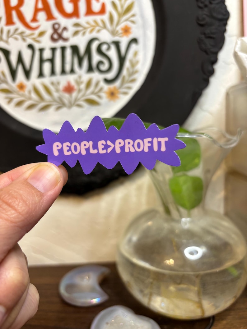 May include: A purple sticker with a jagged edge, displaying the text "PEOPLE>PROFIT" in pink. The sticker is held in front of a blurred background featuring a decorative plate with the words "RAGE & WHIMSY".