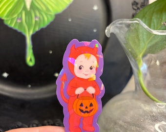 Puede incluir: Una pegatina con un bebé disfrazado de diablo, sosteniendo una linterna de calabaza. El bebé lleva un traje rojo con cuernos y una cola. La pegatina tiene un borde morado y un efecto holográfico. Una polilla verde y un plato negro en el fondo.
