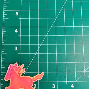 May include: A sticker featuring a stylized horse with a fiery mane and tail. The horse is primarily red and orange, with a yellow outline. The sticker is placed on a green cutting mat with white grid lines.