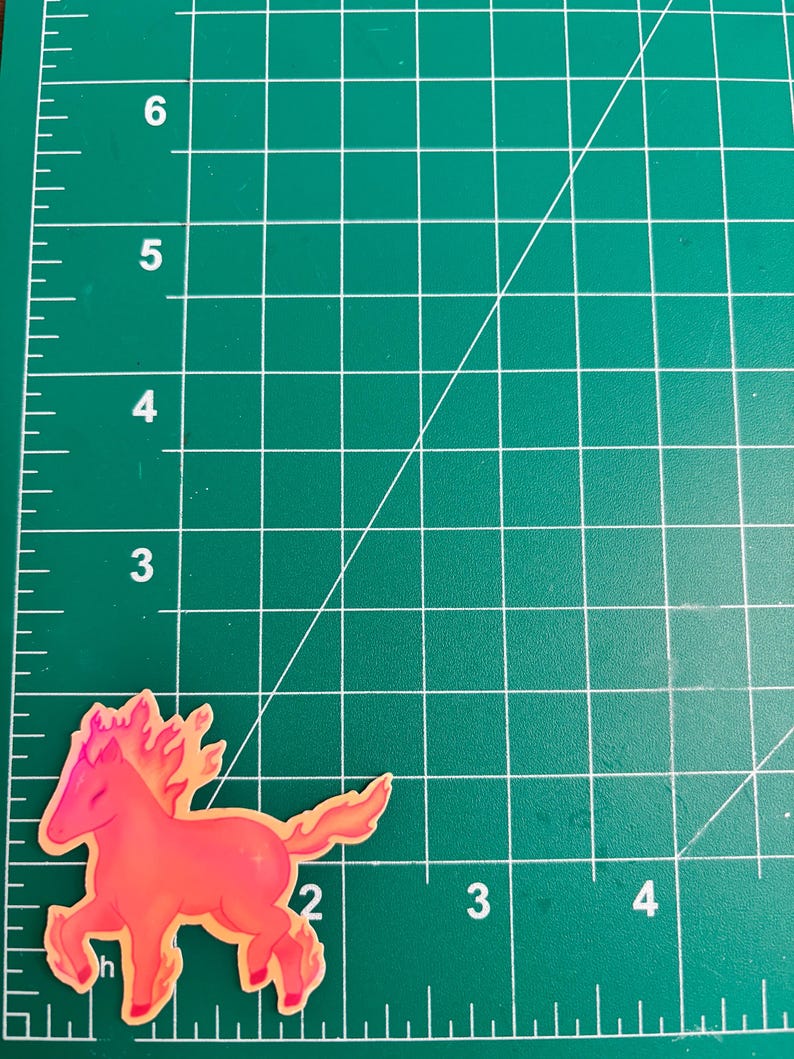 May include: A sticker featuring a stylized horse with a fiery mane and tail. The horse is primarily red and orange, with a yellow outline. The sticker is placed on a green cutting mat with white grid lines.