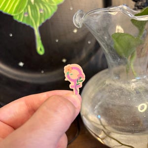 May include: A small, pink and yellow sticker in the shape of the female symbol, decorated with flowers. The sticker is held in a person's hand. In the background, a green and white moth is painted on a black plate.