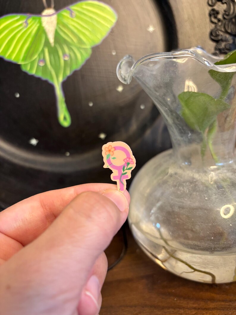 May include: A small, pink and yellow sticker in the shape of the female symbol, decorated with flowers. The sticker is held in a person's hand. In the background, a green and white moth is painted on a black plate.