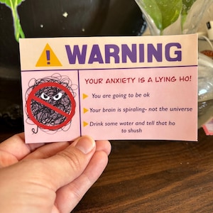 May include: A pink card with the word "WARNING" in purple and yellow. The card has a graphic of a scribbled ball with a red "no" symbol over it. Text on the card reads "YOUR ANXIETY IS A LYING HO!" and other supportive statements.