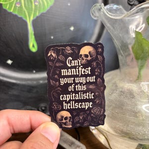 May include: A dark sticker with the text "Can't manifest your way out of this capitalistic hellscape" and illustrations of skulls and roses. The sticker is held by a hand.