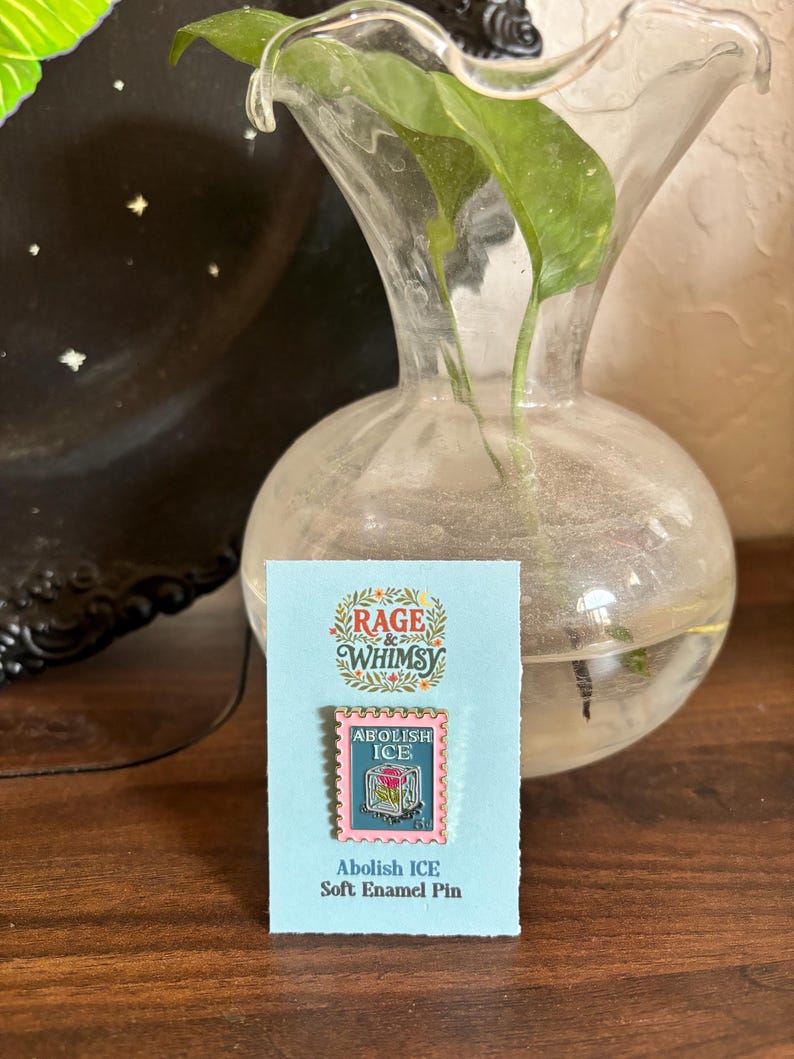 May include: A soft enamel pin on a light blue card. The pin is a pink and teal postage stamp design with the words "ABOLISH ICE" and "RAGE & WHIMSY". A clear glass vase with green leaves is in the background.