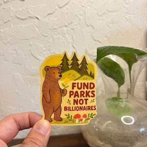 Könnte beinhalten: Ein Aufkleber mit einem braunen Bären, der eine Blume hält, mit dem Text "FUND PARKS NOT BILLIONAIRES". Der Aufkleber hat einen gelben Rand mit Illustrationen von Bäumen, Pilzen und Blumen. Der Bär steht vor einer Waldszene.
