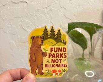 May include: A sticker featuring a brown bear holding a flower, with the text "FUND PARKS NOT BILLIONAIRES." The sticker has a yellow border with illustrations of trees, mushrooms, and flowers. The bear is standing in front of a forest scene.