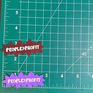 May include: Two die-cut stickers with the text "PEOPLE>PROFIT". One sticker is brown with white text, the other is purple with white text. The stickers are on a green cutting mat with grid lines and measurements in inches.