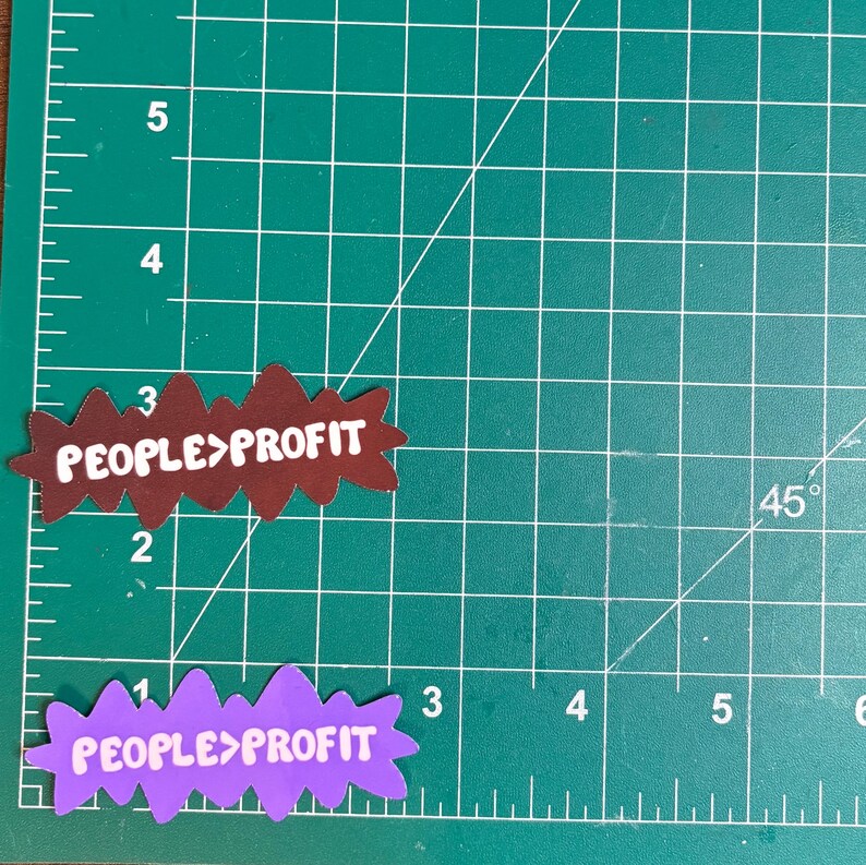 May include: Two die-cut stickers with the text "PEOPLE>PROFIT". One sticker is brown with white text, the other is purple with white text. The stickers are on a green cutting mat with grid lines and measurements in inches.