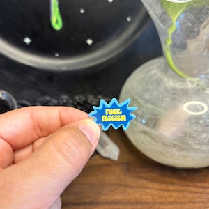 May include: A blue and white pin with the words "FUCK FASCISM" in a speech bubble shape. The pin is held in a hand, with a blurred background featuring a glass vase and a dark, speckled surface.