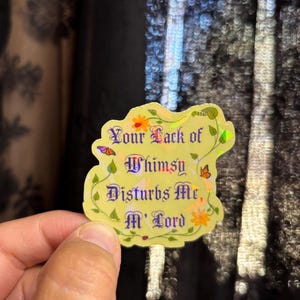 Peut inclure: Un autocollant découpé avec une finition holographique jaune pâle, orné de vignes vertes, de fleurs orange et de petits papillons. Le texte en police gothique violet foncé indique : Your Lack of Whimsy Disturbs Me, M' Lord. Forme irrégulière.