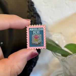 May include: A pink and gold enamel pin in the shape of a postage stamp. The pin features the words "ABOLISH ICE" above a rose in a block of ice. The stamp also has the number "5&cent;".