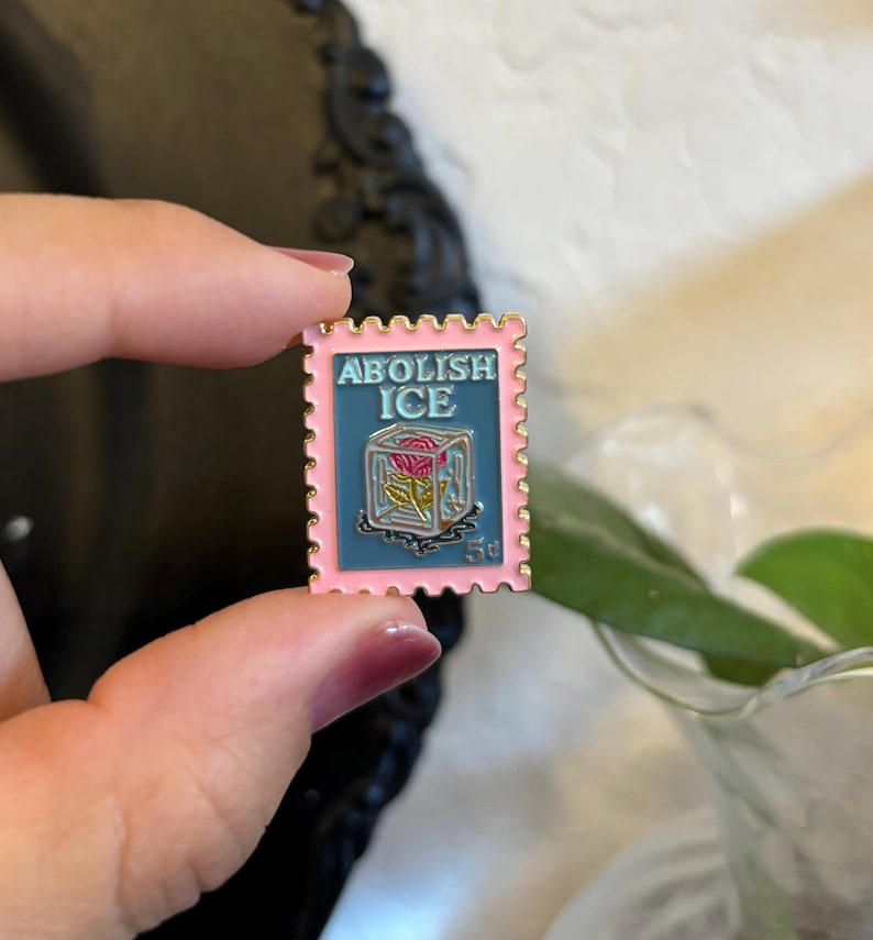 May include: A pink and gold enamel pin in the shape of a postage stamp. The pin features the words "ABOLISH ICE" above a rose in a block of ice. The stamp also has the number "5&cent;".