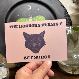Peut inclure: Une carte rose avec le texte "THE HORRORS PERSIST BUT SO DO I" et une illustration de chat noir aux yeux jaunes. La carte est tenue devant une assiette décorative noire.