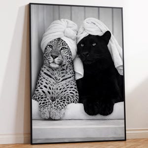 Op de afbeelding: Een zwart-wit ingelijste muurkunstprint met een gevlekte luipaard en een zwarte panter die naast elkaar zitten. Beide dieren hebben witte handdoeken als tulbanden om hun hoofd gewikkeld en rusten hun voorpoten op een witte richel.