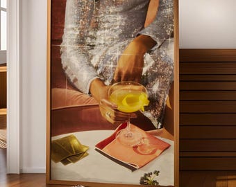 May include: A framed print depicts a person holding a cocktail with a lemon twist. The person wears a sequined silver dress. A gold-colored card, a pink napkin, and a small cluster of berries are on the table.