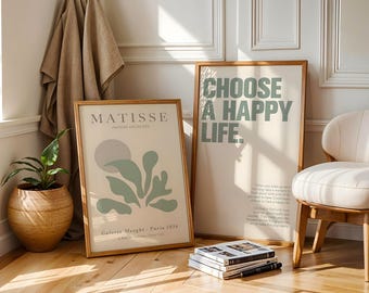 Peut inclure: Deux affiches encadrées avec des cadres en bois sont appuyées contre un mur blanc. L'une dit "CHOOSE A HAPPY LIFE" en lettres vertes. L'autre affiche dit "MATISSE" et présente un motif abstrait vert et gris.