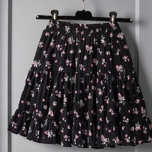 May include: Black tiered skirt with a floral pattern. The skirt features a ruffled hem and an elastic waistband. The flowers are pink and white, and the fabric has a textured appearance. The skirt is hanging on a black hanger.