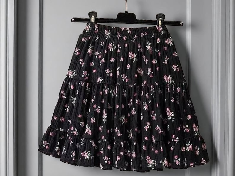 May include: Black tiered skirt with a floral pattern. The skirt features a ruffled hem and an elastic waistband. The flowers are pink and white, and the fabric has a textured appearance. The skirt is hanging on a black hanger.