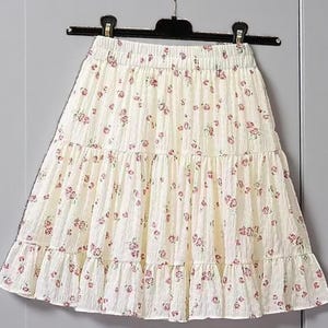May include: A cream-colored, tiered skirt with a floral pattern of small pink flowers. The skirt is hanging on a black hanger against a neutral background. The fabric appears to be lightweight.