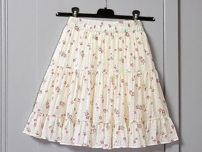 May include: A cream-colored, tiered skirt with a floral pattern of small pink flowers. The skirt is hanging on a black hanger against a neutral background. The fabric appears to be lightweight.