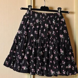 May include: Black tiered skirt with a floral pattern. The skirt is hanging on a black hanger. The skirt has a ruffled hem and is made of a textured fabric. The floral pattern features pink and white flowers.