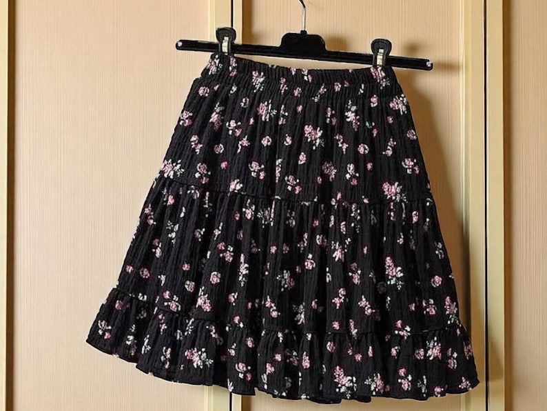 May include: Black tiered skirt with a floral pattern. The skirt is hanging on a black hanger. The skirt has a ruffled hem and is made of a textured fabric. The floral pattern features pink and white flowers.