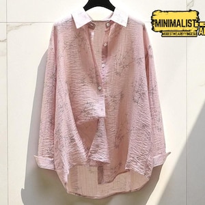May include: A light pink button-down shirt with a white collar and cuffs, hanging on a black hanger. The shirt has a subtle, textured pattern and a loose fit. The text "MINIMALIST APPAREL" is in a yellow and black box.