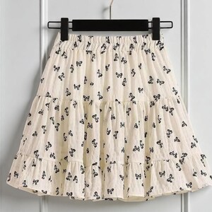 May include: A cream-colored skirt with a tiered design, adorned with a repeating pattern of black butterfly motifs. The skirt is hanging on a black hanger against a white background. The skirt has a ruffled hem.